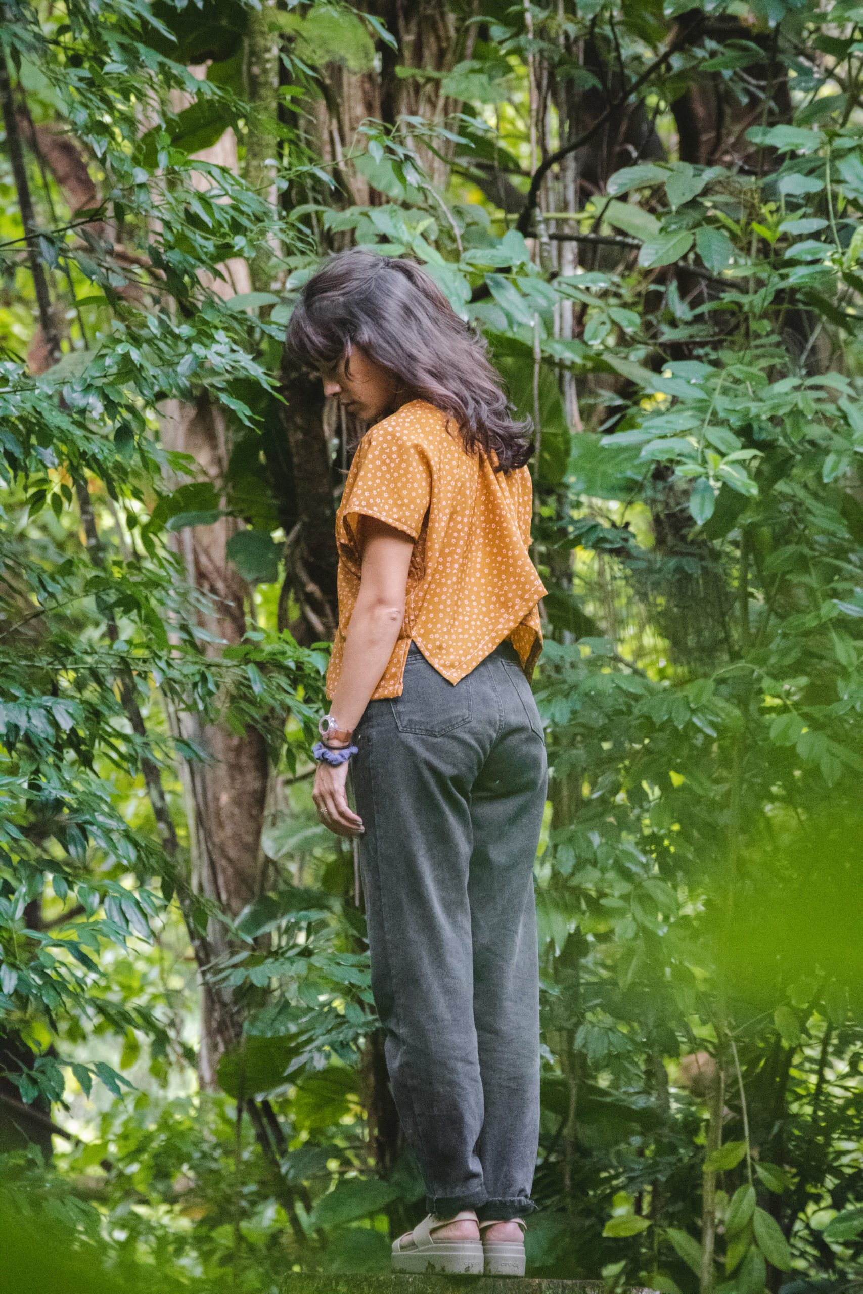 Woman in an orange top and black jeans looks down with a forested background