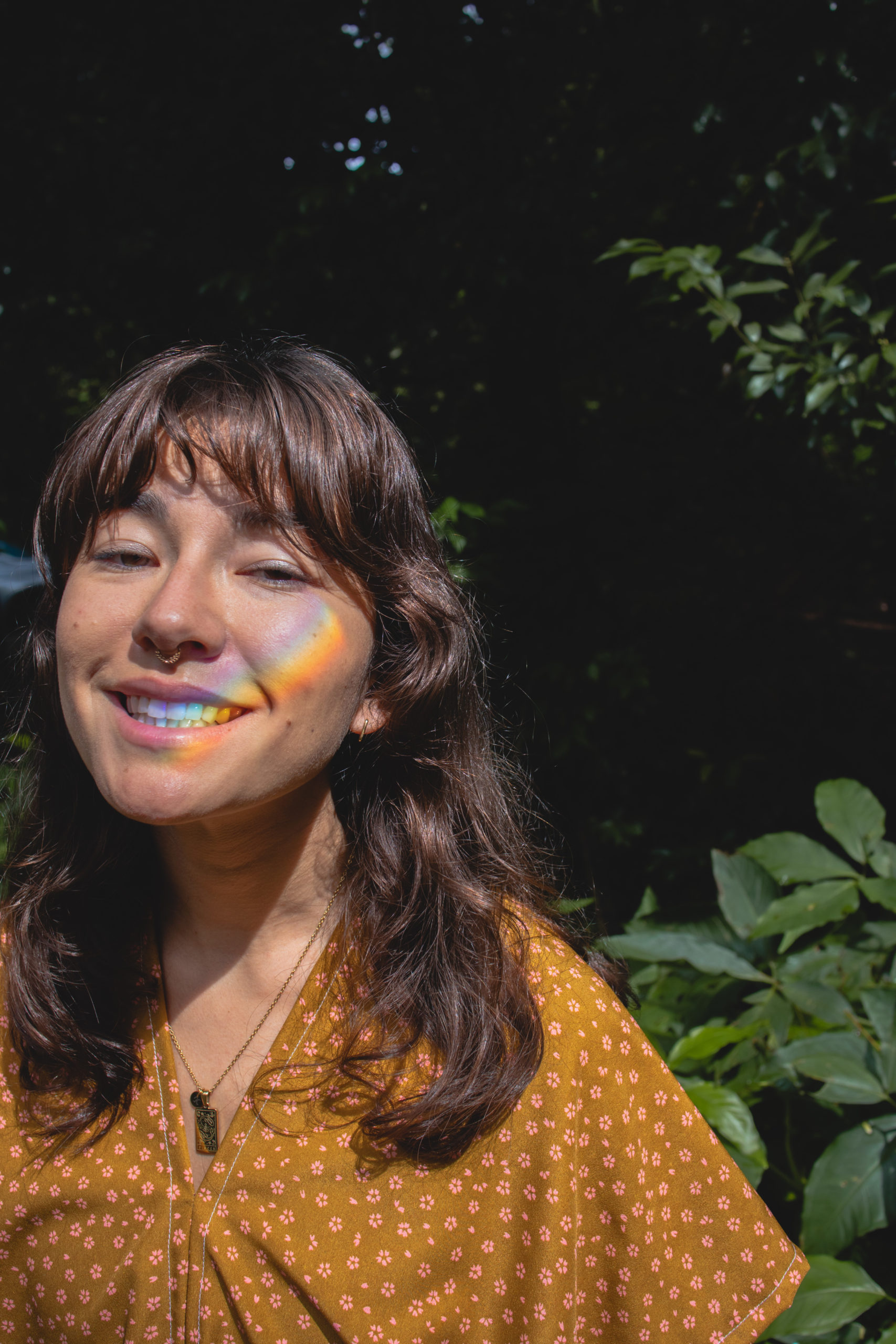 Woman smiles at the camera with a forest in the background and rainbow light on her face