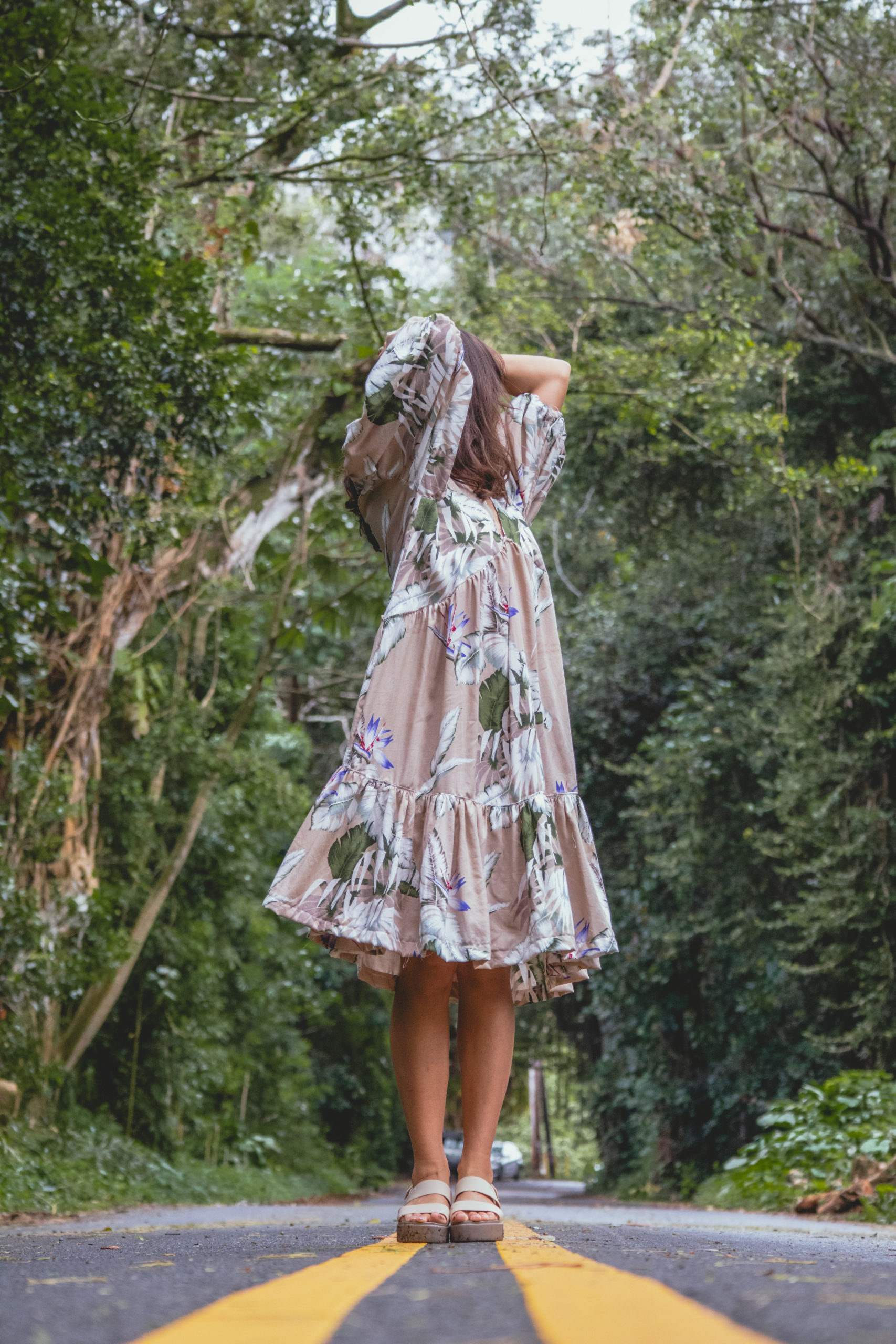 Woman in a beige dress holds her hair and looks away from the camera with forest in the background.